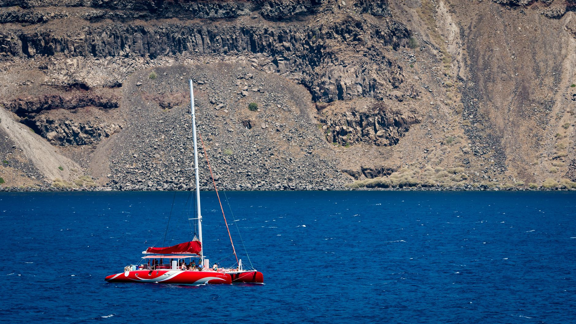 Red sailing boat on the Aegean below Santorini’s volcanic cliffs